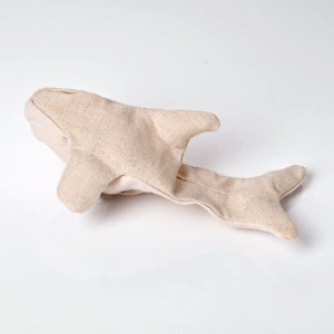 May include: A beige fabric shark toy with fins and a tail. The toy is made of a textured material and is lying on a white surface. The shark has a light beige color.