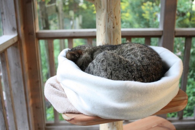 Image shows a grey and white stripped cat curled up in our white organic cotton sleep sack.  The cat and organic bed are perched on a wooden cat tree in front of a window looking out on a green landscape.  Natural Organic Cat Beds by Purrfectplay.