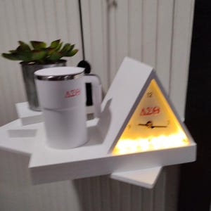 May include: A white mug and a triangular clock with the letters "AZB" in red are displayed on a white shelf. The clock has a yellow illuminated face. A small potted plant is also on the shelf.