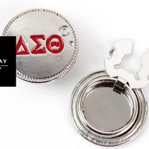 May include: Silver-toned phone ring with a red and silver Greek letter design. The ring features the Greek letters ΔΣΘ. The ring is round with a textured edge and a hinged, foldable stand. The logo "MON'TAY COLLECTION MOCHA MANE" is in the image.