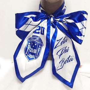 May include: A white and blue satin scarf with the text "Zeta Phi Beta" and a crest with the text "ZOB 1920". The scarf has a blue border and a diamond pattern.
