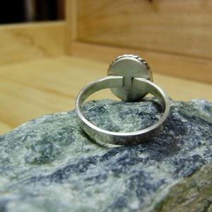 May include: A silver ring with a round, smooth stone set in the centre. The ring is on a grey rock.