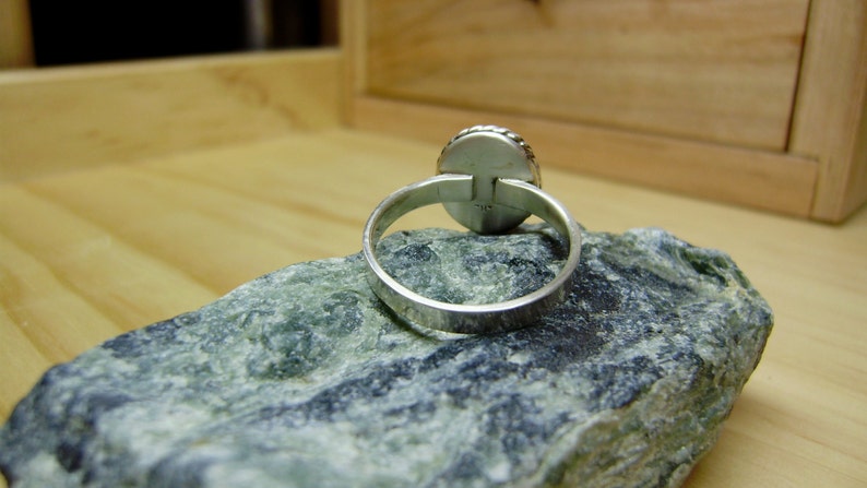 May include: A silver ring with a round, smooth stone set in the centre. The ring is on a grey rock.