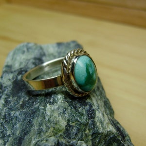 May include: A silver ring with a green gemstone set in a silver bezel. The ring is on a grey rock.