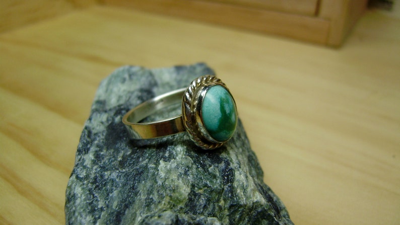 May include: A silver ring with a green gemstone set in a silver bezel. The ring is on a grey rock.