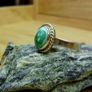 May include: A silver ring with a green gemstone set in a gold bezel. The ring is on a grey rock.