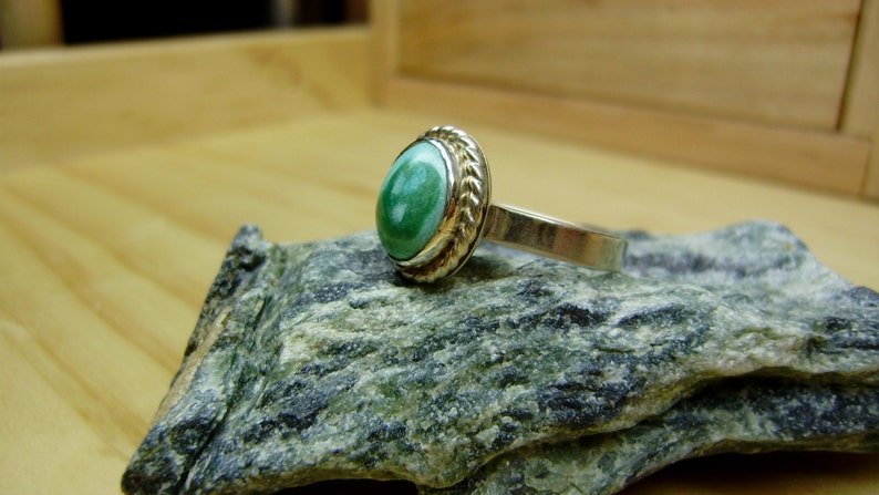 May include: A silver ring with a green gemstone set in a gold bezel. The ring is on a grey rock.