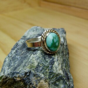 May include: A silver ring with a green oval gemstone set in a silver bezel. The ring is on a grey rock.