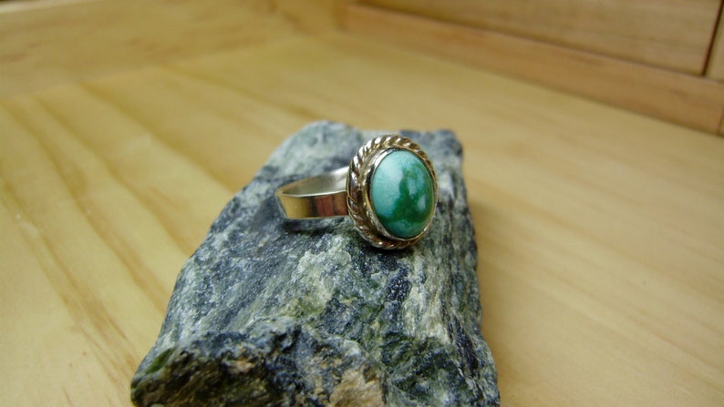 May include: A silver ring with a green oval gemstone set in a silver bezel. The ring is on a grey rock.