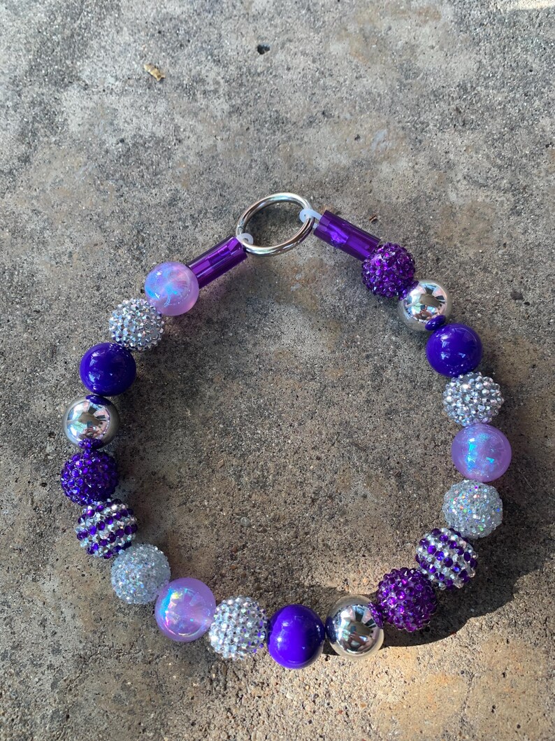 May include: A purple and silver beaded dog collar with a silver clasp. The beads are various sizes and shapes, including round, square, and star-shaped.