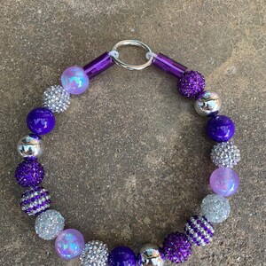 May include: A purple and silver beaded necklace with a silver ring clasp. The beads are various sizes and shapes, including round, oval, and faceted.