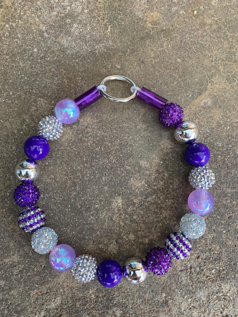 May include: A purple and silver beaded necklace with a silver ring clasp. The beads are various sizes and shapes, including round, oval, and faceted.