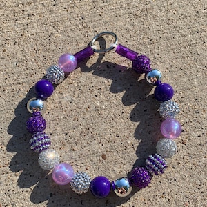 May include: A purple and silver beaded necklace with a silver clasp. The beads are various sizes and shapes, including round, oval, and square. Some of the beads are covered in glitter.
