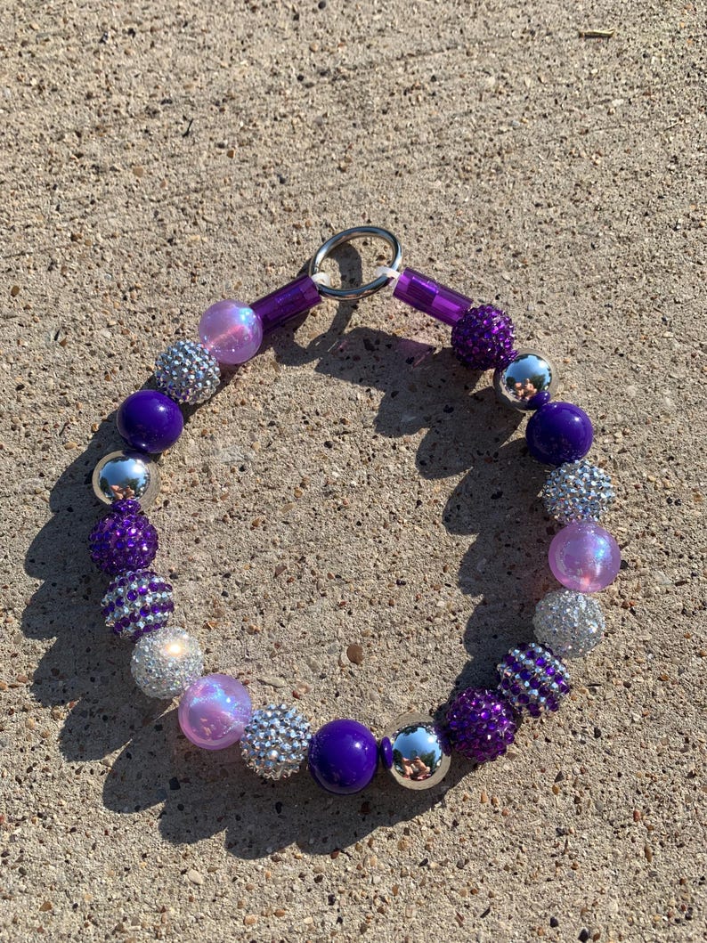 May include: A purple and silver beaded necklace with a silver clasp. The beads are various sizes and shapes, including round, oval, and square. Some of the beads are covered in glitter.