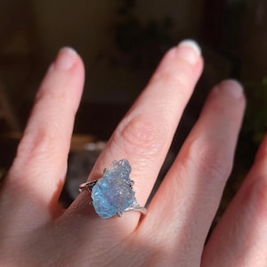 May include: A light blue aquamarine ring. The gemstone is a rough, irregular shape with a light blue hue. The ring's band is silver-colored and simple, with a prong setting holding the stone. The ring is displayed on a hand, with the focus on the gemstone.