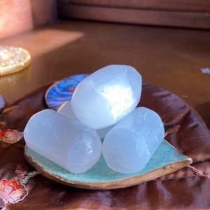 May include: Three white selenite stones, smooth and polished, are arranged on a small, blue and green ceramic dish. The dish is sitting on a brown, satin-like pouch with intricate embroidery.