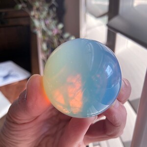 May include: A translucent, spherical crystal ball with a gradient of colors, including blue, yellow, and orange. The crystal ball is held in a hand, and the light reflects off the surface, creating a glowing effect. The crystal ball is a decorative item.
