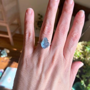May include: A ring featuring a large, raw, light blue gemstone set in a silver-toned metal band. The stone has an irregular, textured surface and is flanked by two small, clear, round-cut accent stones on either side of the central setting.
