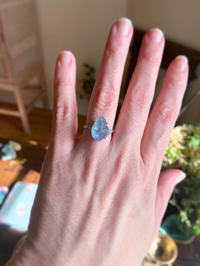 May include: A ring featuring a large, raw, light blue gemstone set in a silver-toned metal band. The stone has an irregular, textured surface and is flanked by two small, clear, round-cut accent stones on either side of the central setting.