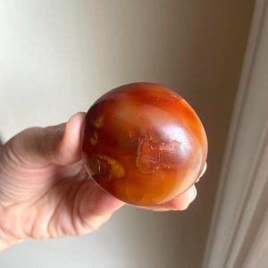 May include: A polished, spherical carnelian stone featuring a smooth, glossy surface with deep orange, reddish-brown, and translucent amber color variations. The stone displays natural internal inclusions and swirling patterns throughout its round form.