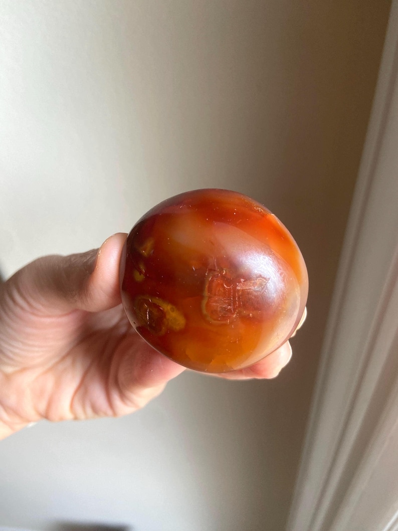 May include: A polished, spherical carnelian stone featuring a smooth, glossy surface with deep orange, reddish-brown, and translucent amber color variations. The stone displays natural internal inclusions and swirling patterns throughout its round form.