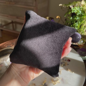 May include: A small, dark gray, square-shaped pillow made of soft fabric. The pillow is held in a hand, with a blurred background featuring plants and decorative items. The pillow's texture appears smooth and plush.