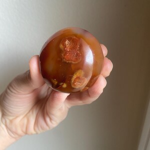 May include: A polished, spherical carnelian stone featuring a translucent, deep reddish-orange hue. The surface displays natural, darker inclusions and circular, amber-colored patterns embedded within the smooth, rounded mineral structure.