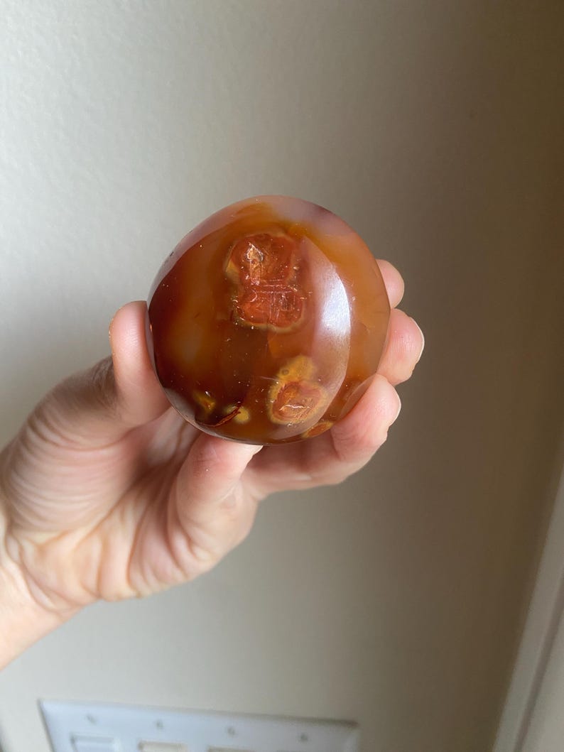May include: A polished, spherical carnelian stone featuring a translucent, deep reddish-orange hue. The surface displays natural, darker inclusions and circular, amber-colored patterns embedded within the smooth, rounded mineral structure.
