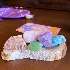 May include: Assortment of colorful crystals including rose quartz, amethyst, and turquoise, displayed on a natural stone base. A tarot card and a purple pouch are in the background. The crystals vary in size and shape.