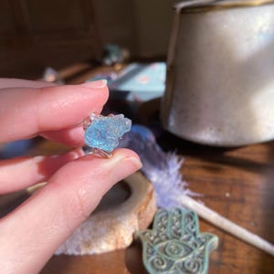 May include: A ring featuring a raw, light blue, translucent gemstone set in a silver-toned metal band. The stone has an irregular, jagged, and crystalline texture with visible facets that catch the light, creating a bright, icy blue appearance.