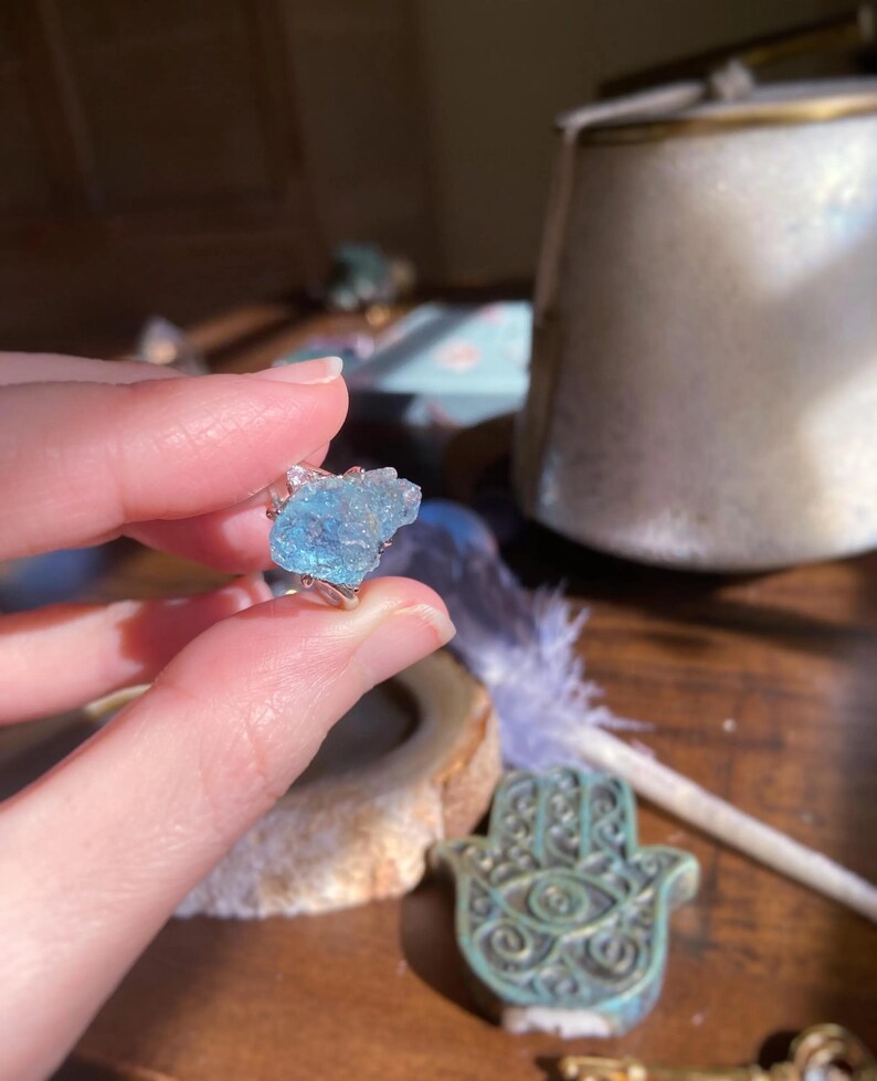 May include: A ring featuring a raw, light blue, translucent gemstone set in a silver-toned metal band. The stone has an irregular, jagged, and crystalline texture with visible facets that catch the light, creating a bright, icy blue appearance.