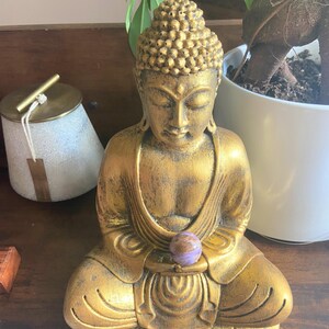 May include: A gold-painted Buddha statue sits in meditation with a purple and brown sphere in its hands.