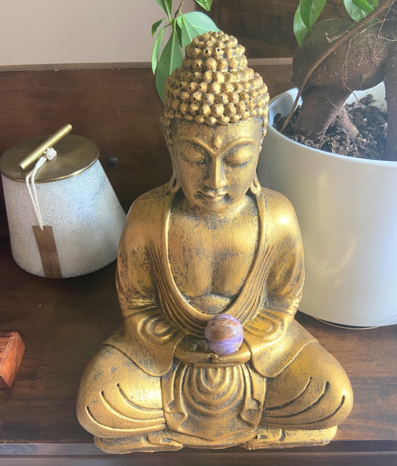 May include: A gold-painted Buddha statue sits in meditation with a purple and brown sphere in its hands.