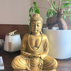 May include: A gold-colored Buddha statue sitting in a meditative pose with hands in lap. The statue has a detailed face and intricate carvings on the robe.