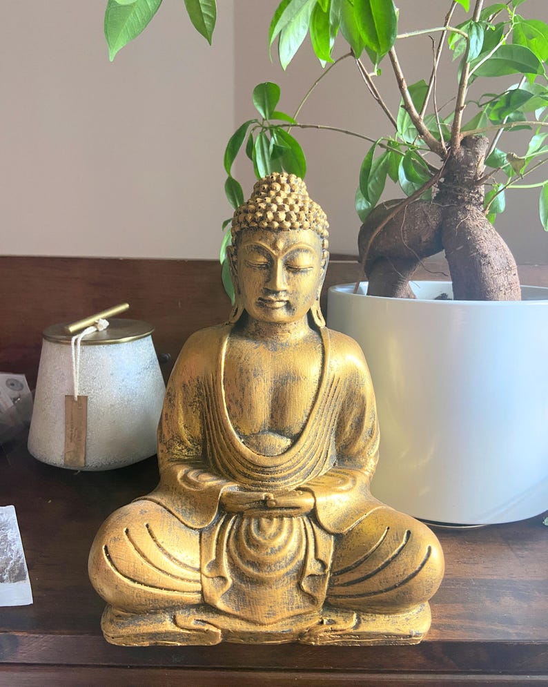 May include: A gold-colored Buddha statue sitting in a meditative pose with hands in lap. The statue has a detailed face and intricate carvings on the robe.