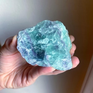May include: A hand holding a rough-cut fluorite crystal. The crystal displays a mix of blue, green, and purple hues. The stone has a natural, irregular shape, with visible textures and variations in color.