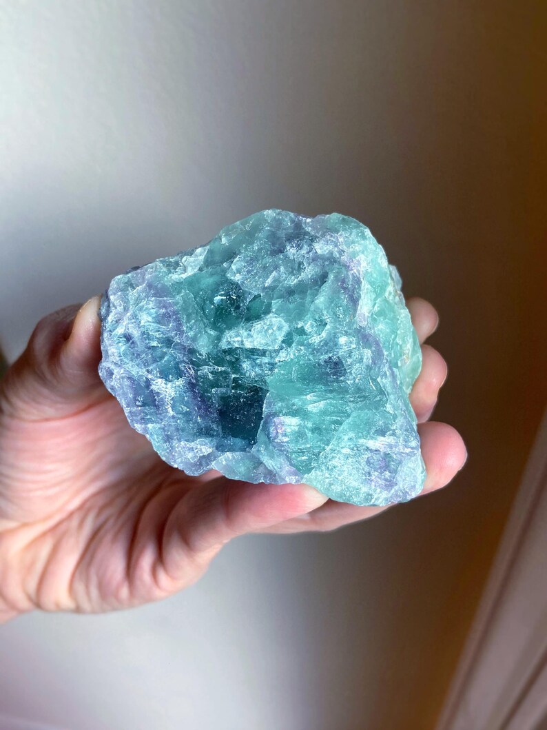 May include: A hand holding a rough-cut fluorite crystal. The crystal displays a mix of blue, green, and purple hues. The stone has a natural, irregular shape, with visible textures and variations in color.