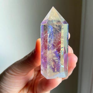 Puede incluir: Una punta de cristal hexagonal transparente con un revestimiento iridiscente que refleja tonos vibrantes de morado, azul y dorado. La piedra pulida presenta una parte superior puntiaguda y un cuerpo facetado con fracturas internas.