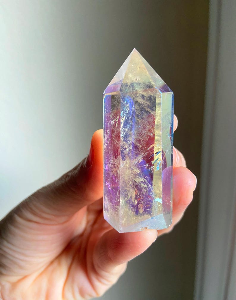 Puede incluir: Una punta de cristal hexagonal transparente con un revestimiento iridiscente que refleja tonos vibrantes de morado, azul y dorado. La piedra pulida presenta una parte superior puntiaguda y un cuerpo facetado con fracturas internas.