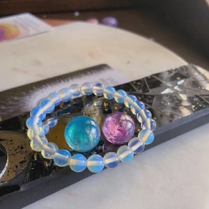 May include: A beaded bracelet with opalescent beads encircles two glass spheres, one teal and one purple. The bracelet and spheres rest on a dark, textured surface, creating a contrast of colors and textures.
