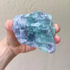 May include: A raw, natural fluorite crystal in shades of blue, green, and purple. The mineral is held in a hand, showcasing its rough, textured surface. The crystal's colors blend together, creating a unique and eye-catching appearance.