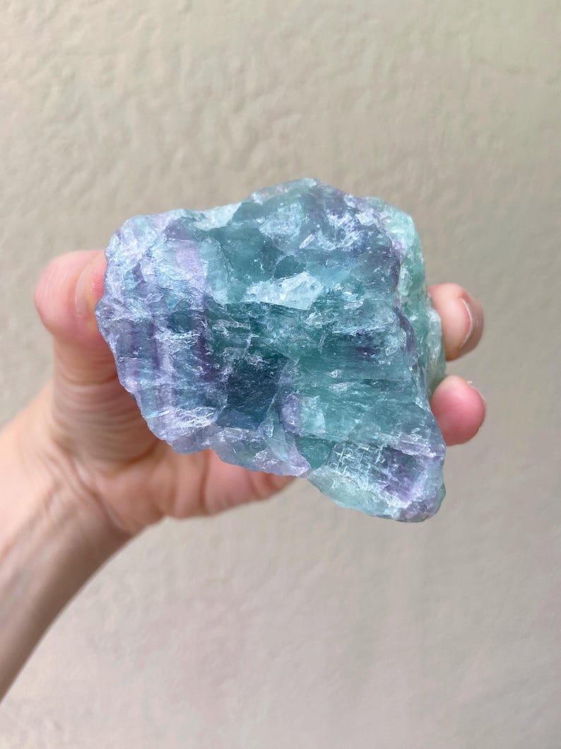 May include: A raw, natural fluorite crystal in shades of blue, green, and purple. The mineral is held in a hand, showcasing its rough, textured surface. The crystal's colors blend together, creating a unique and eye-catching appearance.