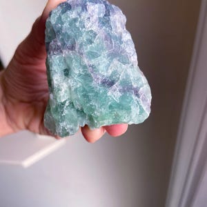 May include: A raw, natural fluorite crystal with a mix of green, blue, and purple hues. The stone has a rough, textured surface and is held in a hand. The crystal's colors blend together, creating a unique and eye-catching appearance.