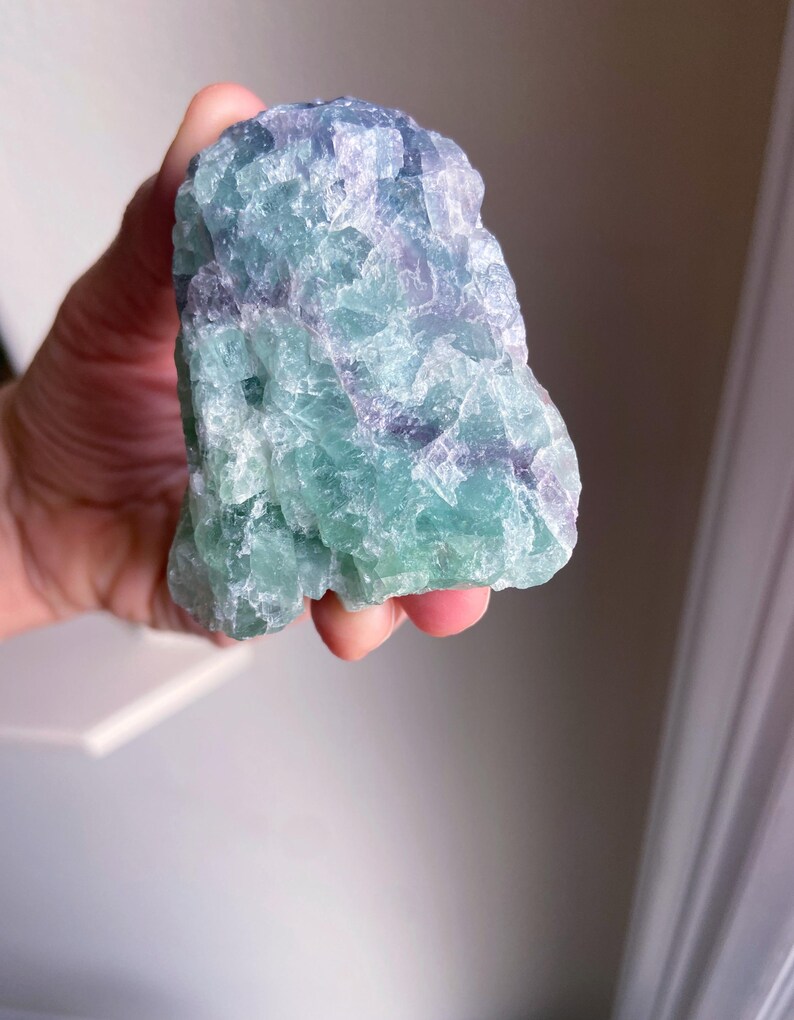 May include: A raw, natural fluorite crystal with a mix of green, blue, and purple hues. The stone has a rough, textured surface and is held in a hand. The crystal's colors blend together, creating a unique and eye-catching appearance.
