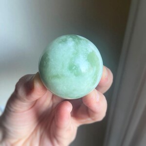 May include: A smooth, spherical jade stone with a pale green hue and white marbling. The polished surface reflects light, showcasing the natural variations in the stone. This decorative sphere is a unique home decor item.
