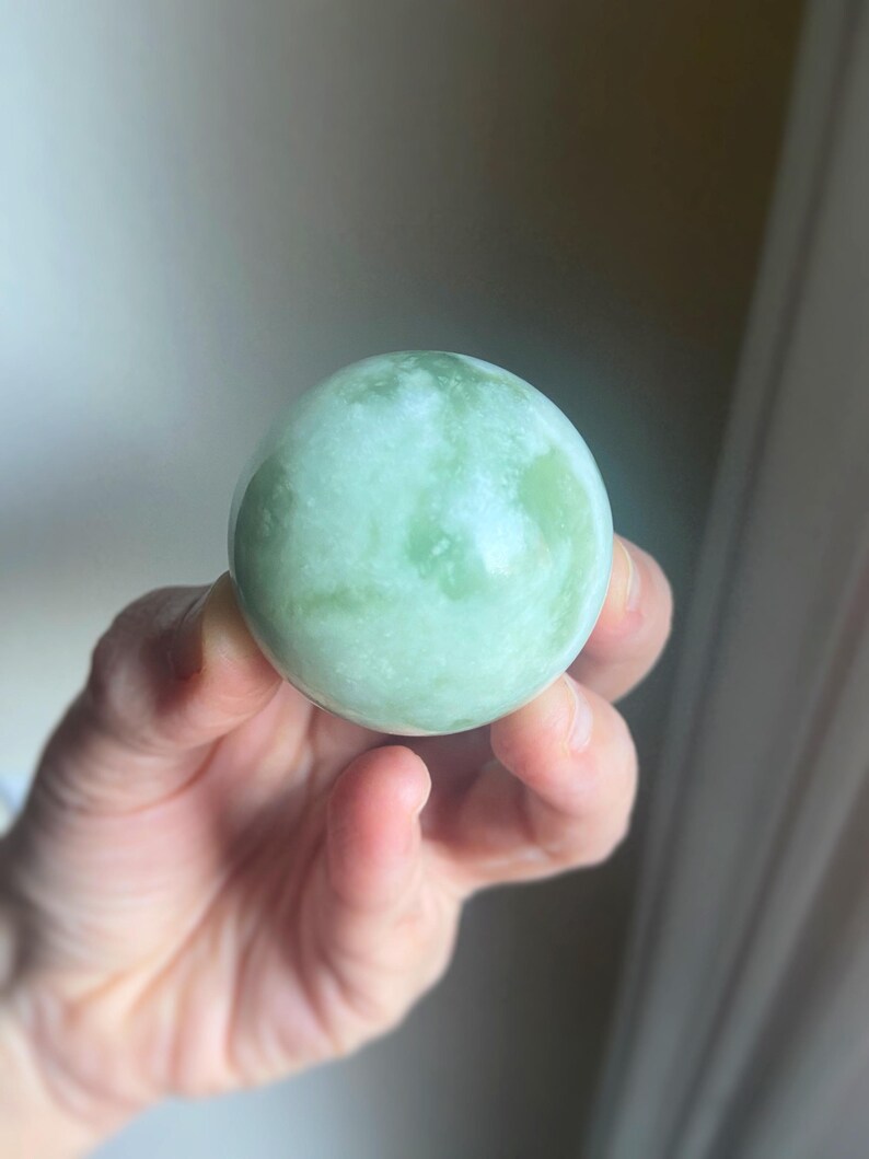 May include: A smooth, spherical jade stone with a pale green hue and white marbling. The polished surface reflects light, showcasing the natural variations in the stone. This decorative sphere is a unique home decor item.