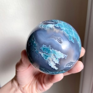 Pu&ograve; includere: Una pietra sferica lucidata con un motivo a spirale di blu, verde acqua e bianco. La sfera &egrave; tenuta in una mano, mostrando la sua superficie liscia e le variazioni naturali di colore. La pietra sembra essere un oggetto decorativo.