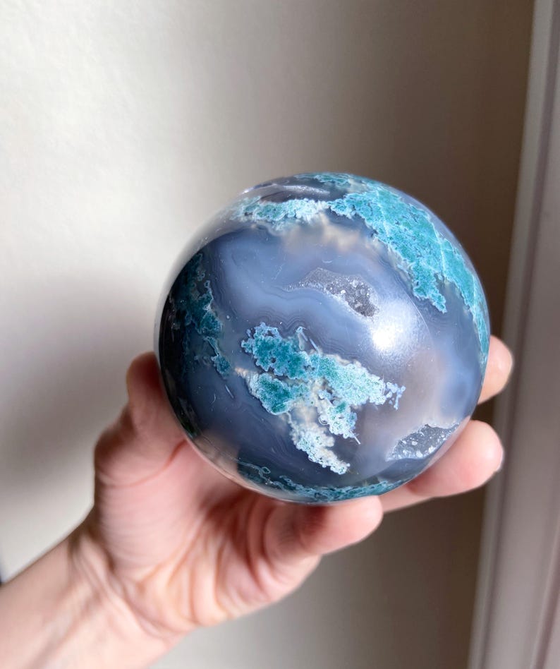 Pu&ograve; includere: Una pietra sferica lucidata con un motivo a spirale di blu, verde acqua e bianco. La sfera &egrave; tenuta in una mano, mostrando la sua superficie liscia e le variazioni naturali di colore. La pietra sembra essere un oggetto decorativo.