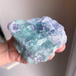 May include: A raw, natural fluorite crystal with a mix of blue, green, and purple hues. The stone has a rough, textured surface and is held in a hand. The crystal is a unique, natural mineral specimen.