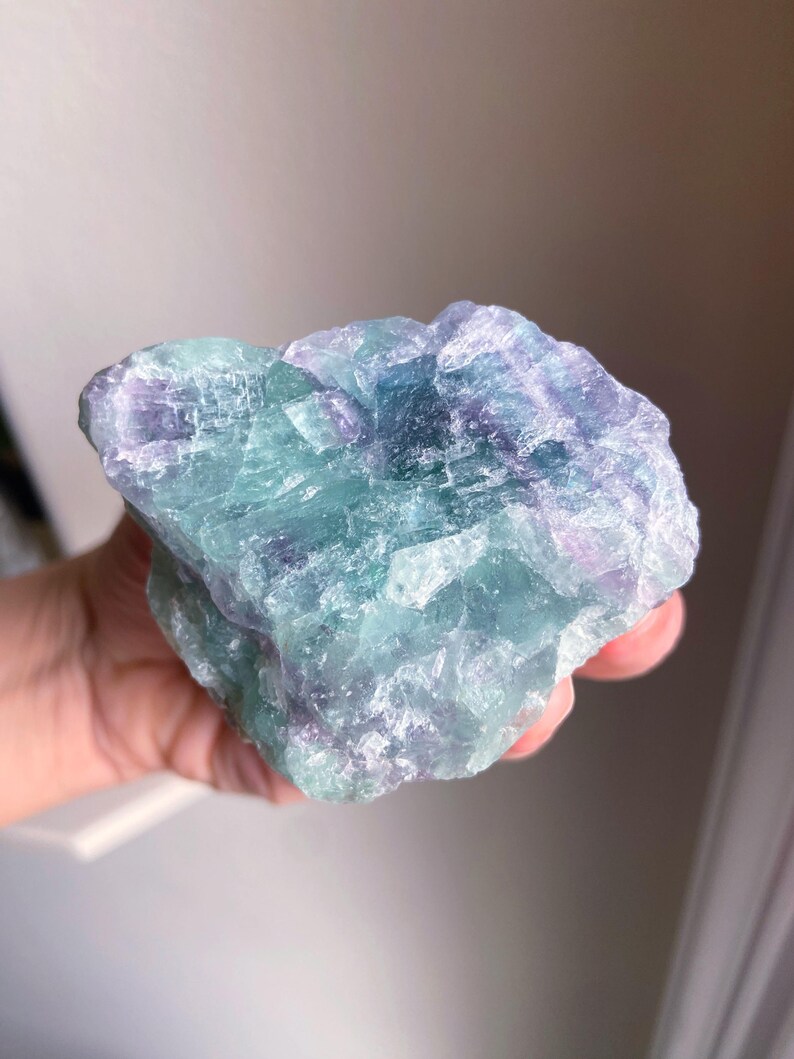 May include: A raw, natural fluorite crystal with a mix of blue, green, and purple hues. The stone has a rough, textured surface and is held in a hand. The crystal is a unique, natural mineral specimen.